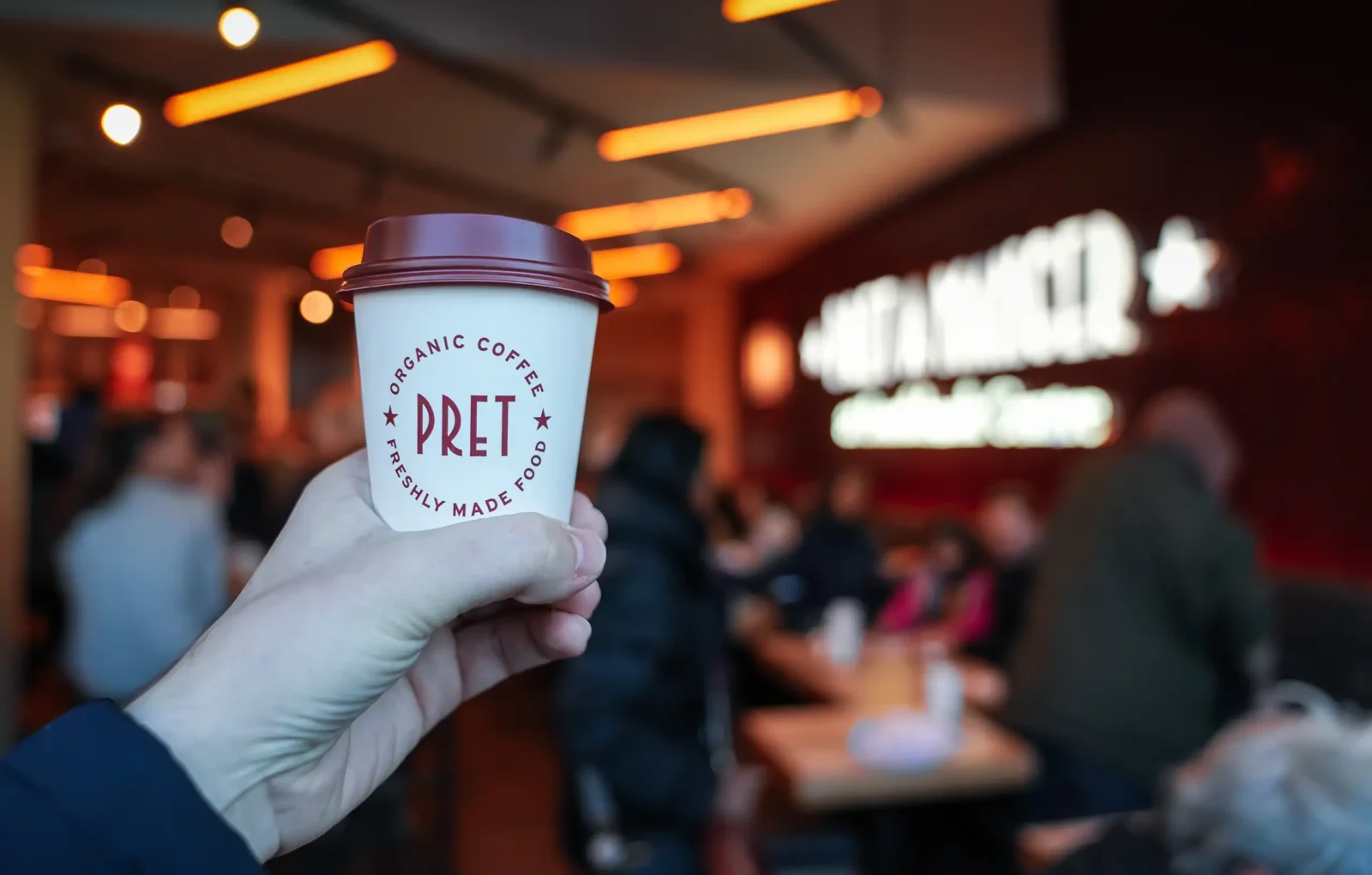 A hand holding up a takeaway coffee cup inside a Pret a Manger.